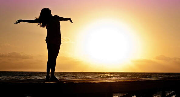 Woman standing on the beach with her arms outstretched looking at the setting yellow sun